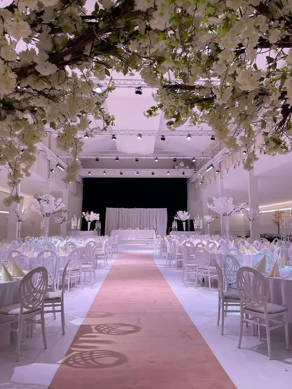 Hochzeit im B2LOFT: Läufer zur Bühne unter blühenden Bäumen, runde Tafeln mit weißen Stühlen